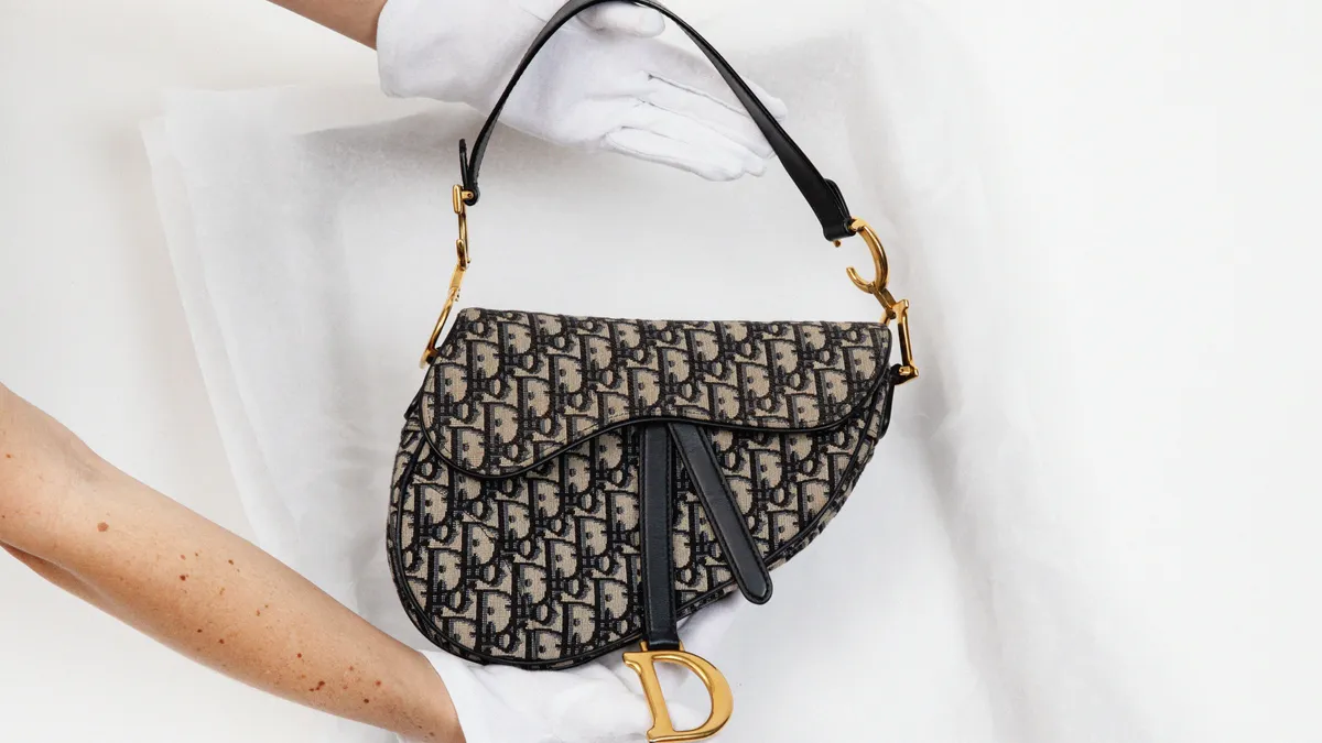 A person wearing white gloves holds up a vintage Dior saddle bag against a white background.