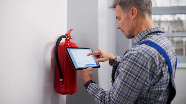 A technician inspects a fire extinguisher while looking at information on a tablet.