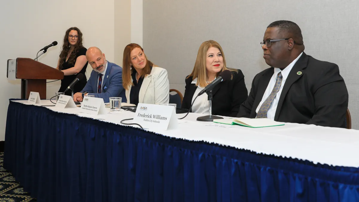 Four people are sitting at a table with microphones and name signs listening and talking. One additional person is standing at a podium.