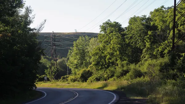 A landfill covered in grass rises behind a line of trees. In the foreground, a two-lane road bends around a curve.