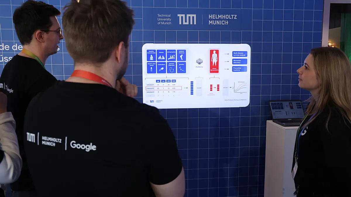 Three people look upon a computer screen built into a blue wall featuring health data information.