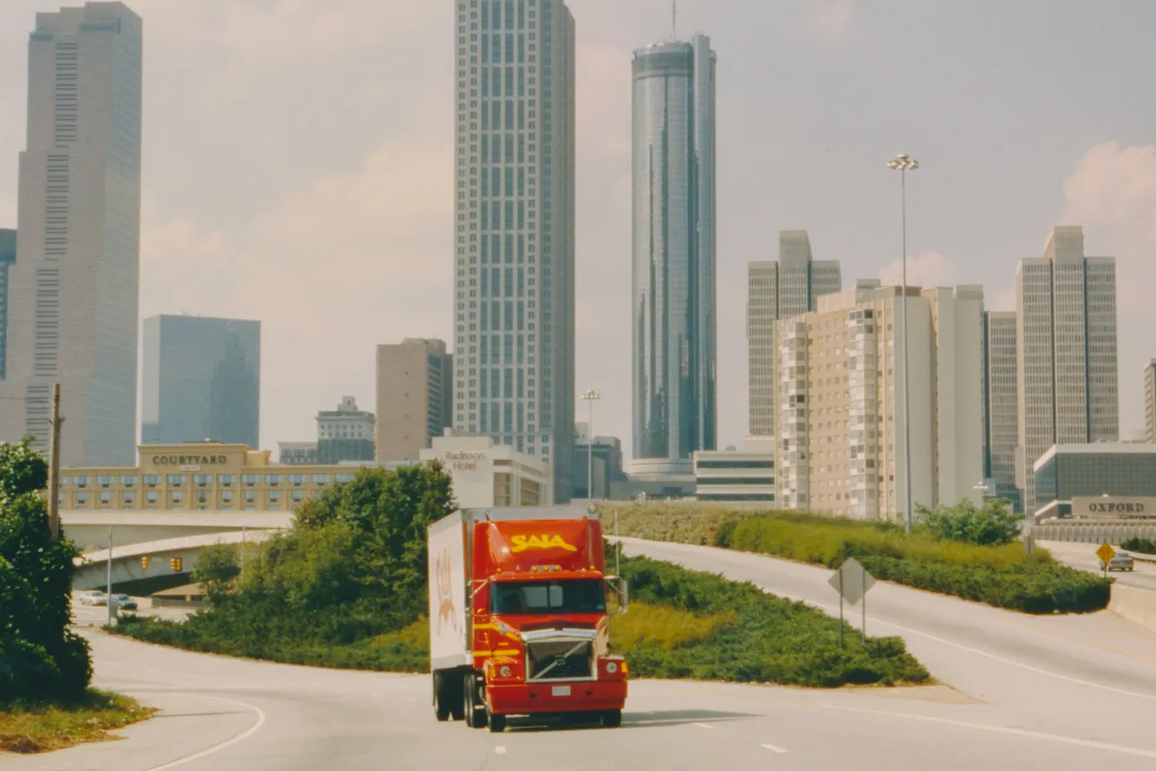 A red Saia Volvo tractor pulls a white Saia trailer on a highway with the Atlanta skyline in the background.