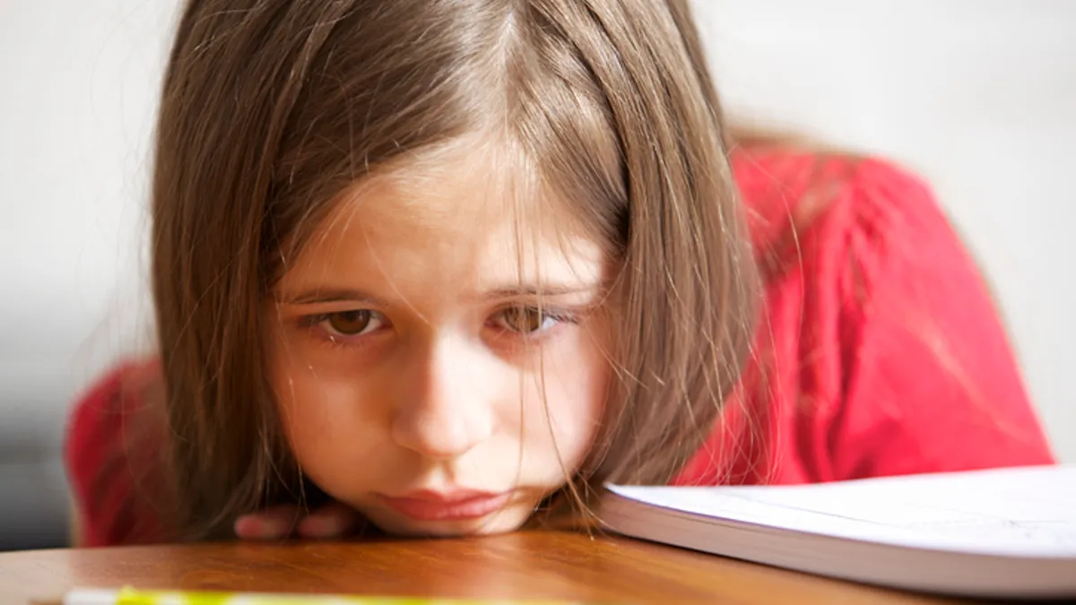 A child has their chin on a desk and looks sad. In front of the child on the desk is a pencil.