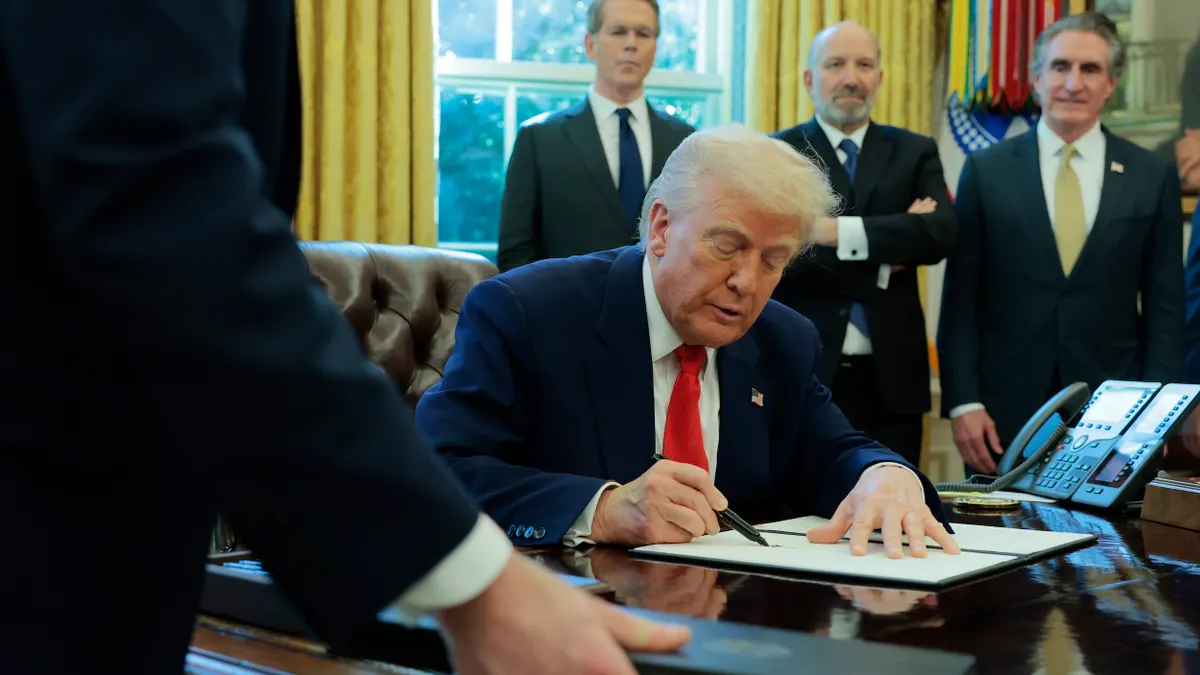President Trump at his Oval Office desk signing an executive order surrounded by men.