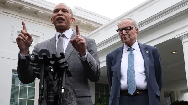 Two men in suits standing in front of the White House. The man on the left is speaking and pointing with both hands.