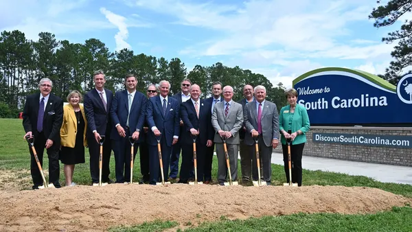 Thirteen people in professional clothing pose with shovels in a pile of dirt.