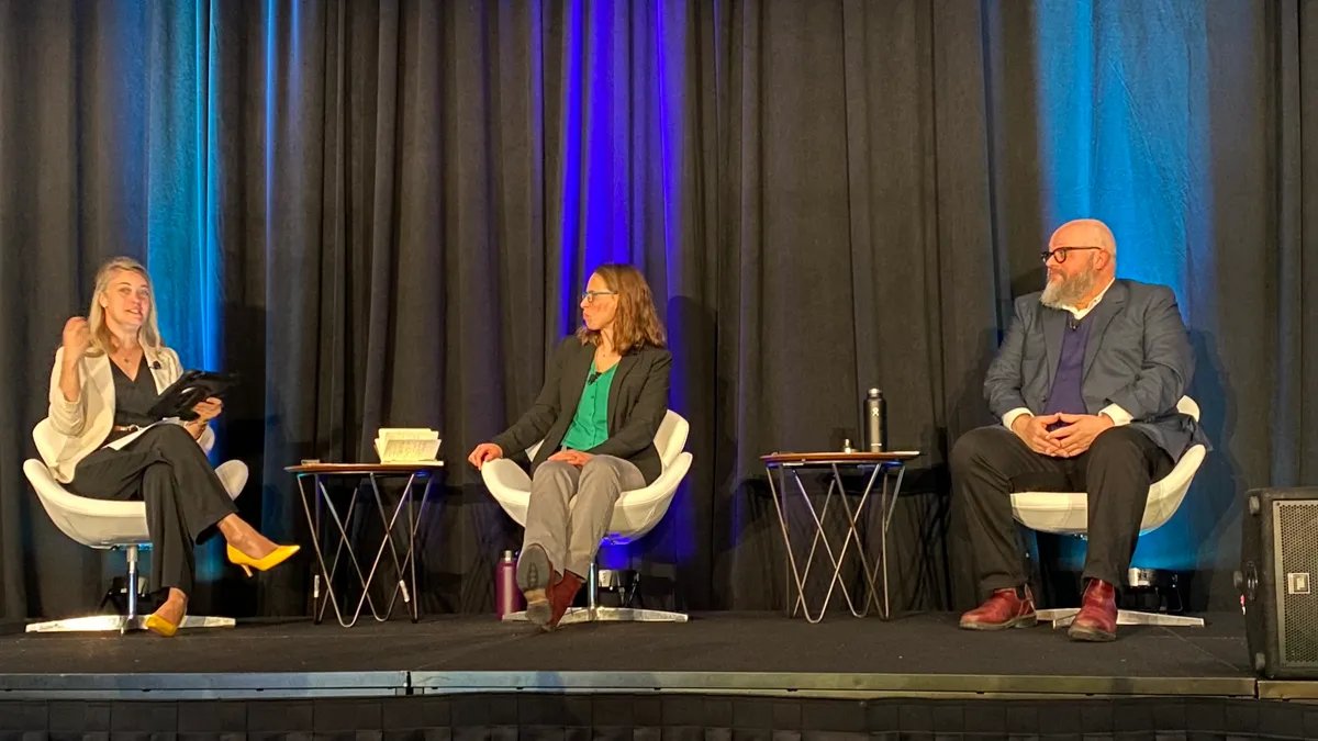 Speakers sit on a stage with a conference presentation in the background.