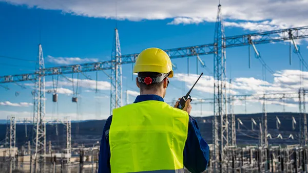 A technician in a fluorescent vest uses a radio beside electrical pylons,
