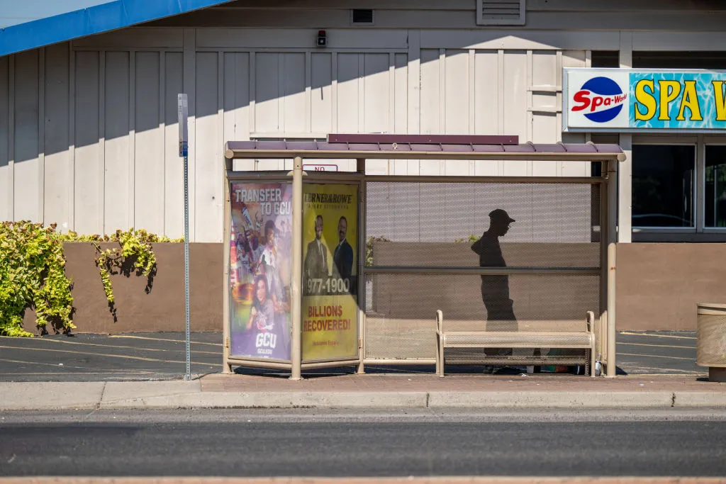 A silhouette of a person wearing a hat under a bus stop on the side of the road. Behind the bus stop is a building.