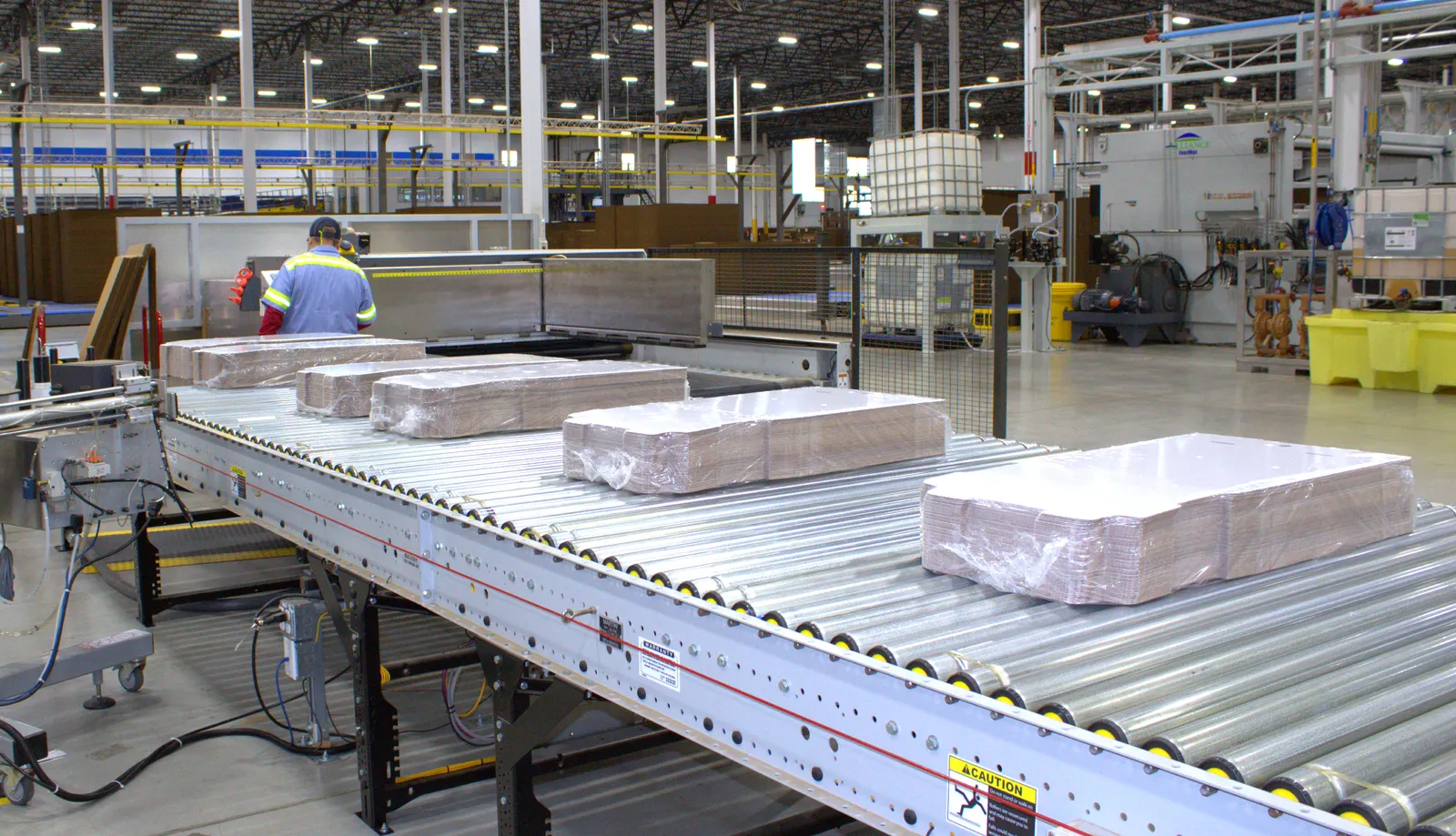 Stacks of plastic-wrapped white boxes move along a conveyor in an industrial facility.