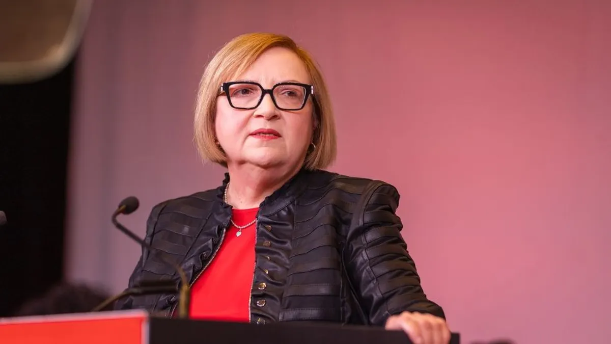 A woman, dressed in black and red, looks to the left as she stands at a black-and-red podium while speaking.