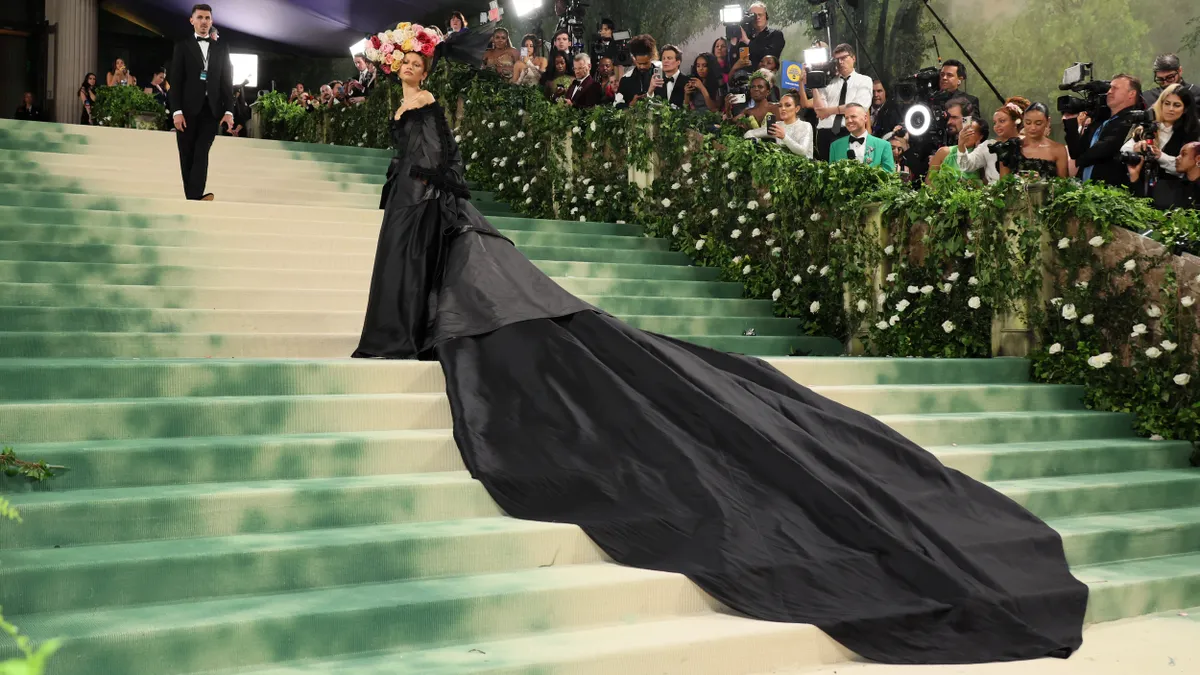 A person stands on steps wearing a large floral hat and a black dress with an extra-long train.