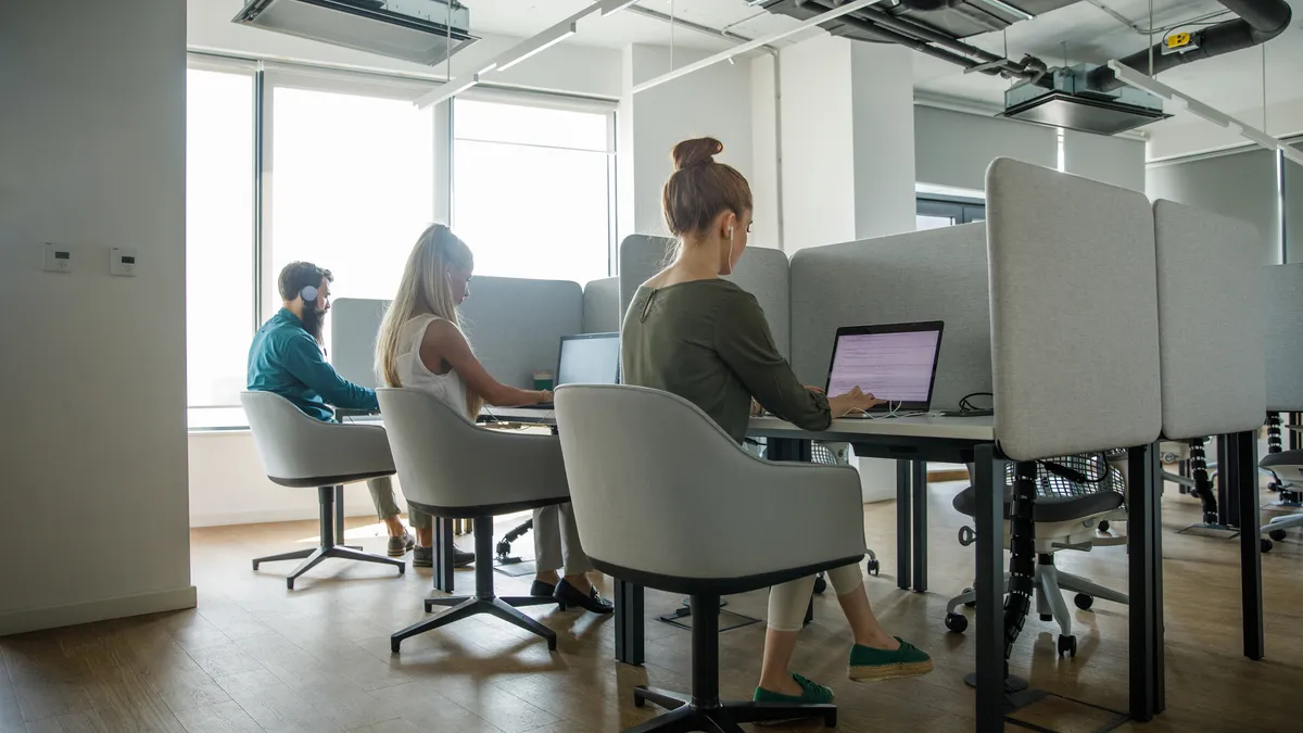 Wide shot of three business people sitting in cubicles, in a communal office space, using laptops.