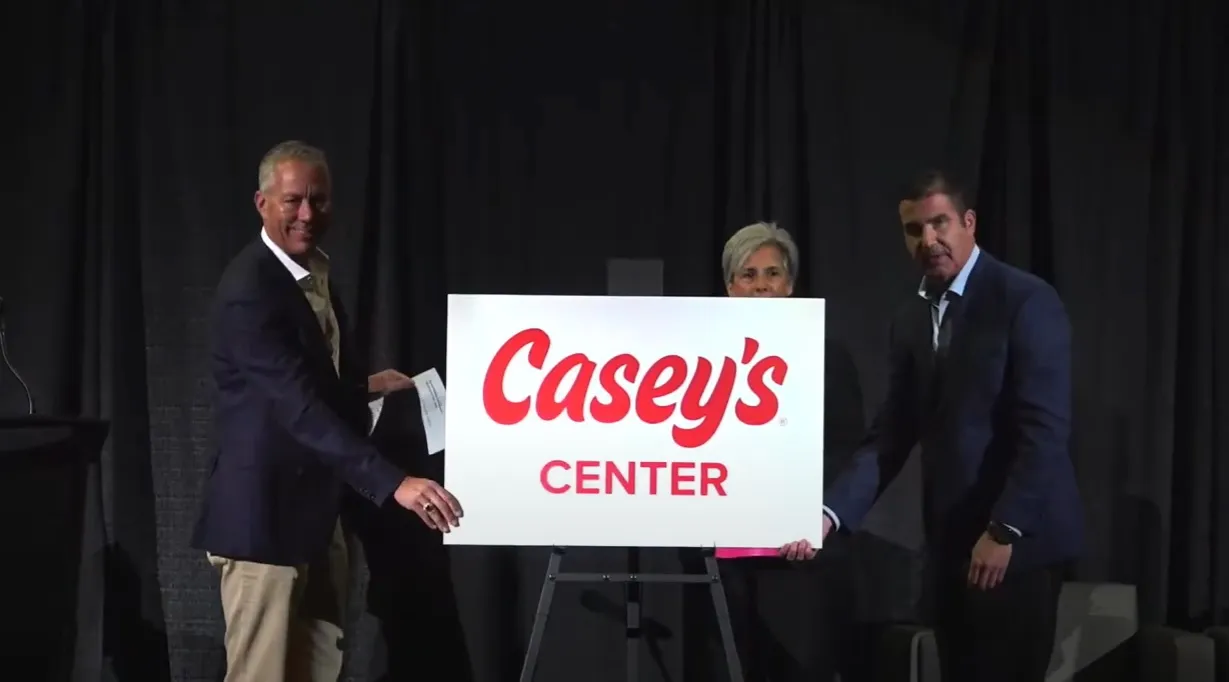 A photo of three people standing on a dark stage around a placard. The placard reads &quot;Casey&#x27;s Center.&quot;