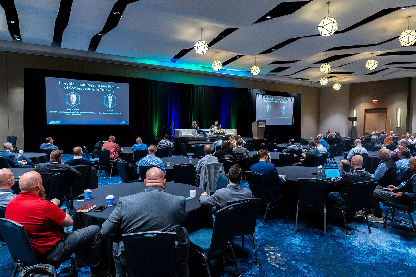 Werner Enterprises SVP and CIO Daragh Mahon talks with NMFTA Chief Operating Officer Joe Ohr about trucking cybersecurity, sitting on a couch on a performance stage with attendees at tables nearby.