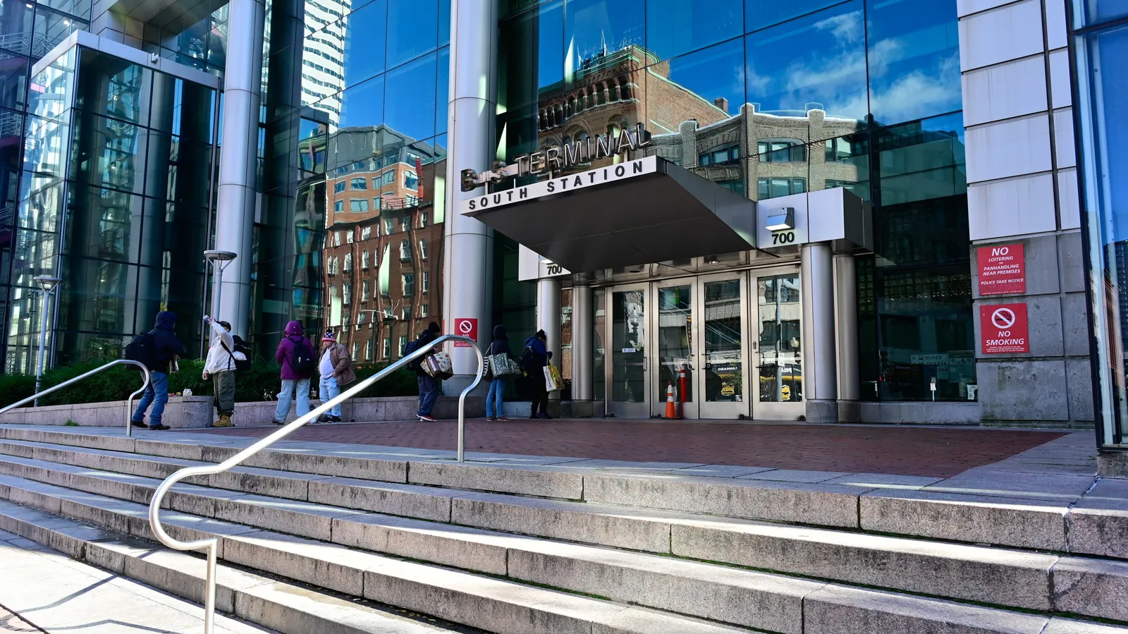 Several people in winter coats stand outside a glass-front building with a sign "Bus Terminal-South Station" over glass doors.