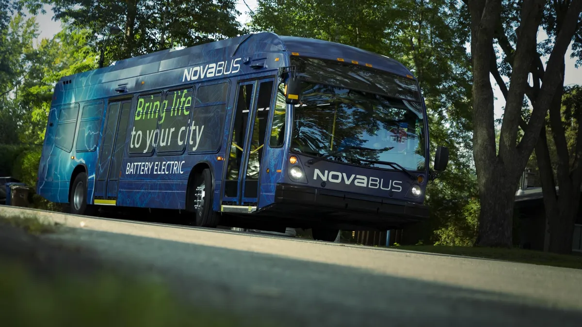 A dark blue electric bus showing a sign on the right side that reads "Nova Bus: Bring life to your city" is on a road under green trees.