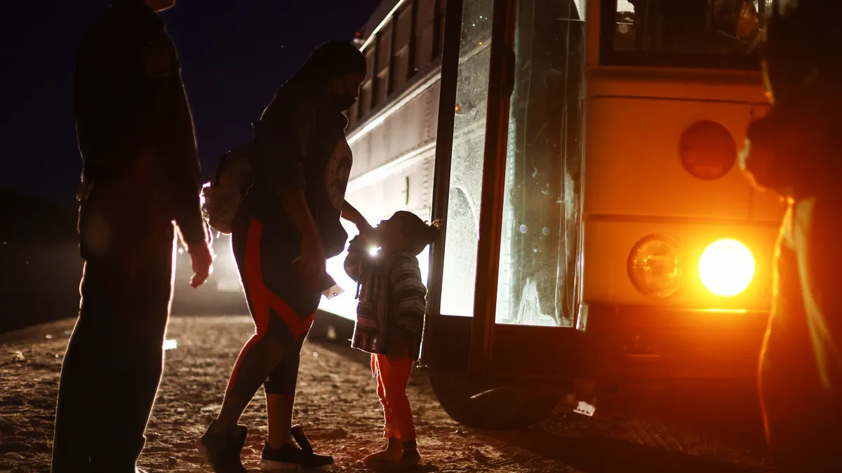 A young child stands outside a bus at night looking up at an adult while another adult stands nearby.