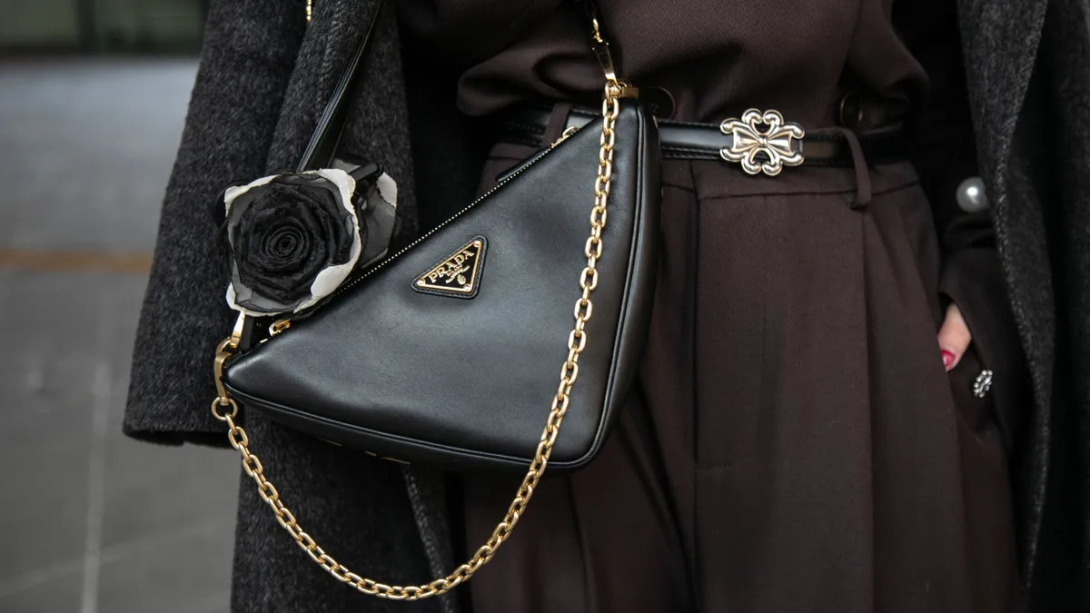 A person stands wearing a black coat and a black Prada triangle bag.