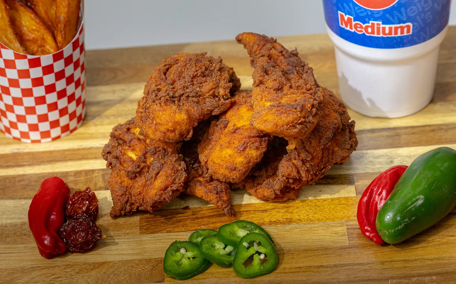 A photo of fried chicken on a wooden surface, surrounded by hot peppers, fries and a cup of soda that says &quot;medium.&quot;