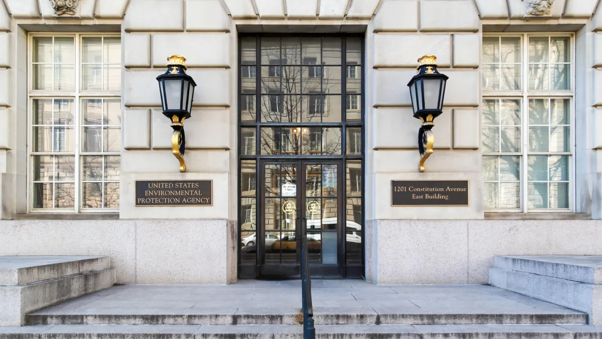 Exterior view of the United States Environmental Protection Agency (EPA) East Building at 1201 Constitution Avenue in Washington, D.C., showing the main entrance with decorative black and gold wall lanterns, large windows, and a central glass door with building reflections.