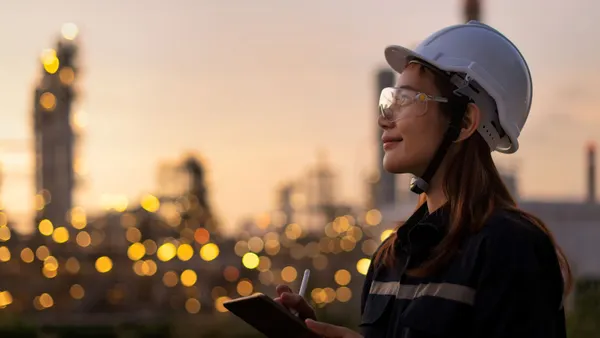 Woman in a hard hat and safety glasses, holding a tablet and stylus, with an industrial facility featuring illuminated structures in the background