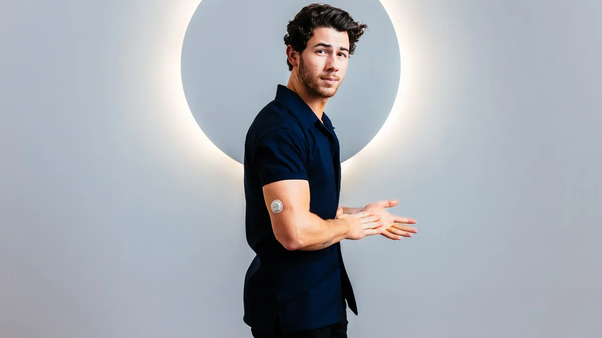 Nick Jonas poses in front of a glowing background, wearing a small, round device on his upper arm