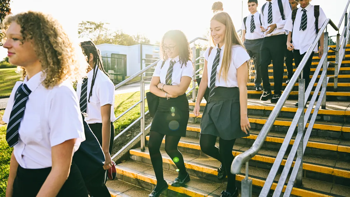 A handful of teen students is walkign down stairs outside on a sunny day. They are wearing uniforms and carrying backpacks.