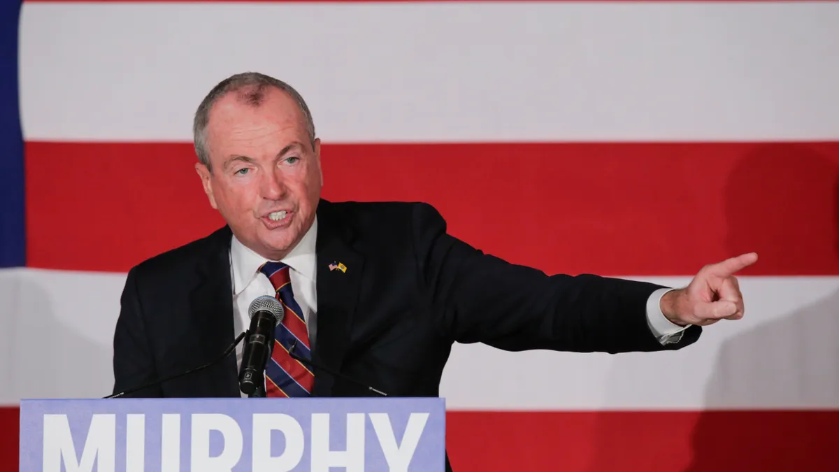 New Jersey Governor Phil Murphy standing at a podium pointing to his left, with flag filling the background.