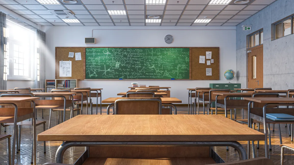 A classroom with no people shows rows of desks with a green chalkboard with writing on the far wall