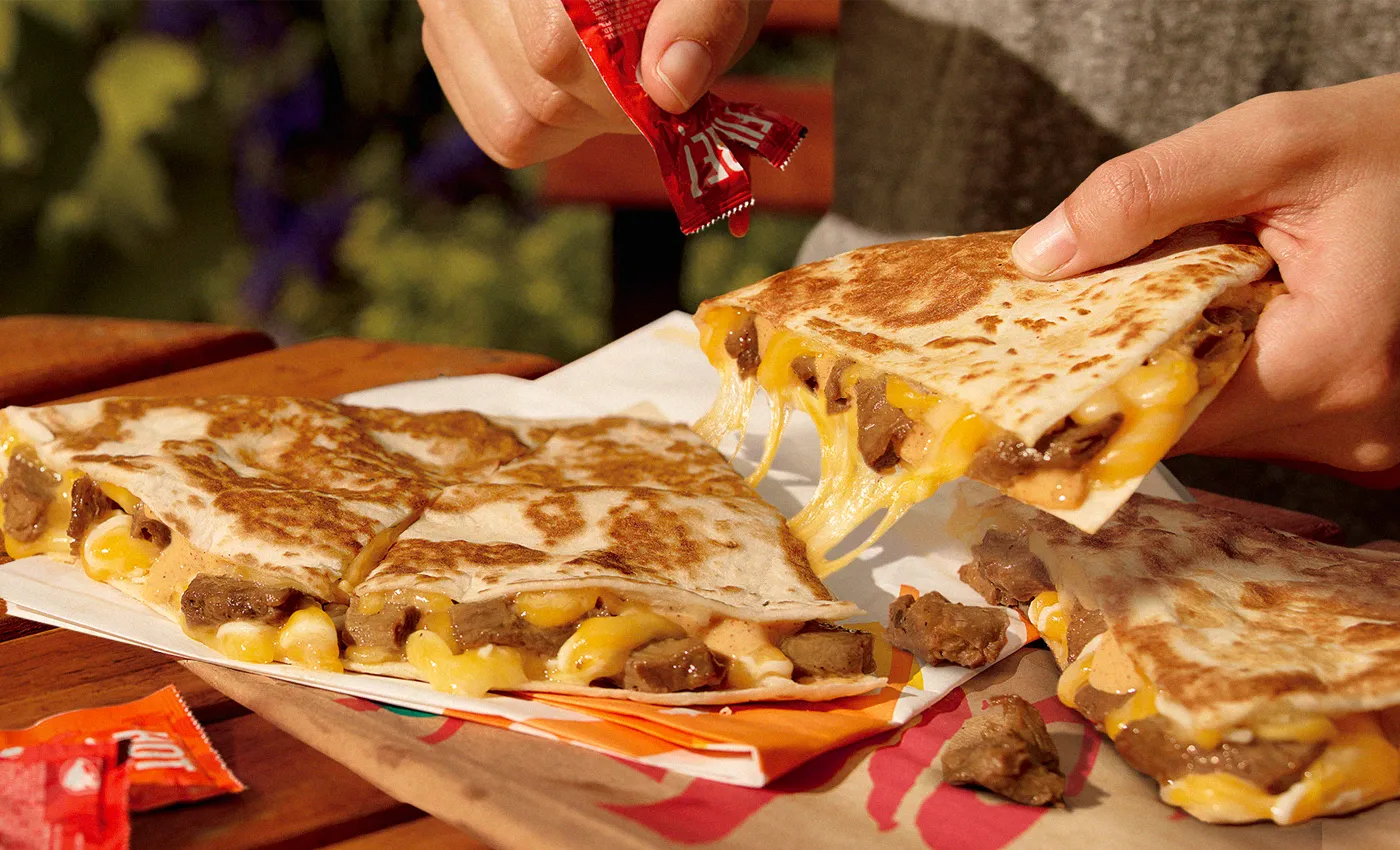 Hands put hot sauce on a large Taco Bell quesadilla made with chunks of plant-based Beyond Carne Asada.