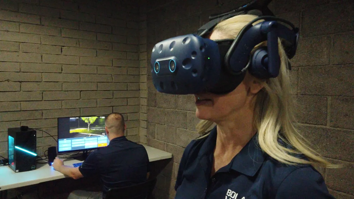 A person is wearing virtual reality goggles as a co-worker looks at a computer.