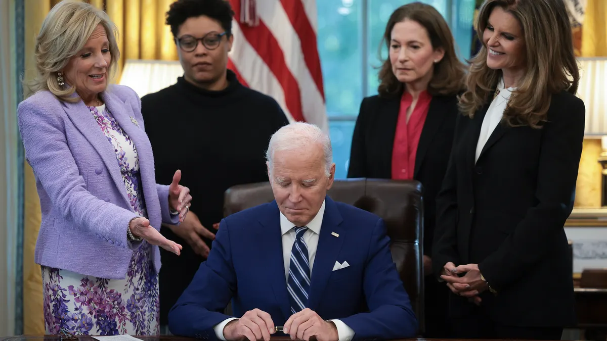 President Joe Biden signs a bill in the Oval Office while First Lady Jill Biden and others look on.