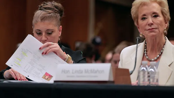 Two people are seated at a table in a room. One person is looking ahead and the other is looking through a pile of papers.