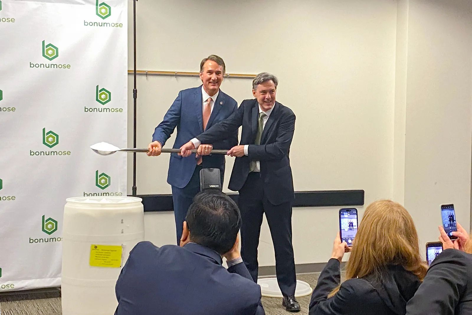 Glenn Youngkin and Ed Rogers hold a large silver spoon of white crystalline tagatose over a white barrel at the front of a conference room with a Bonumose logo backdrop. People take photos in front.