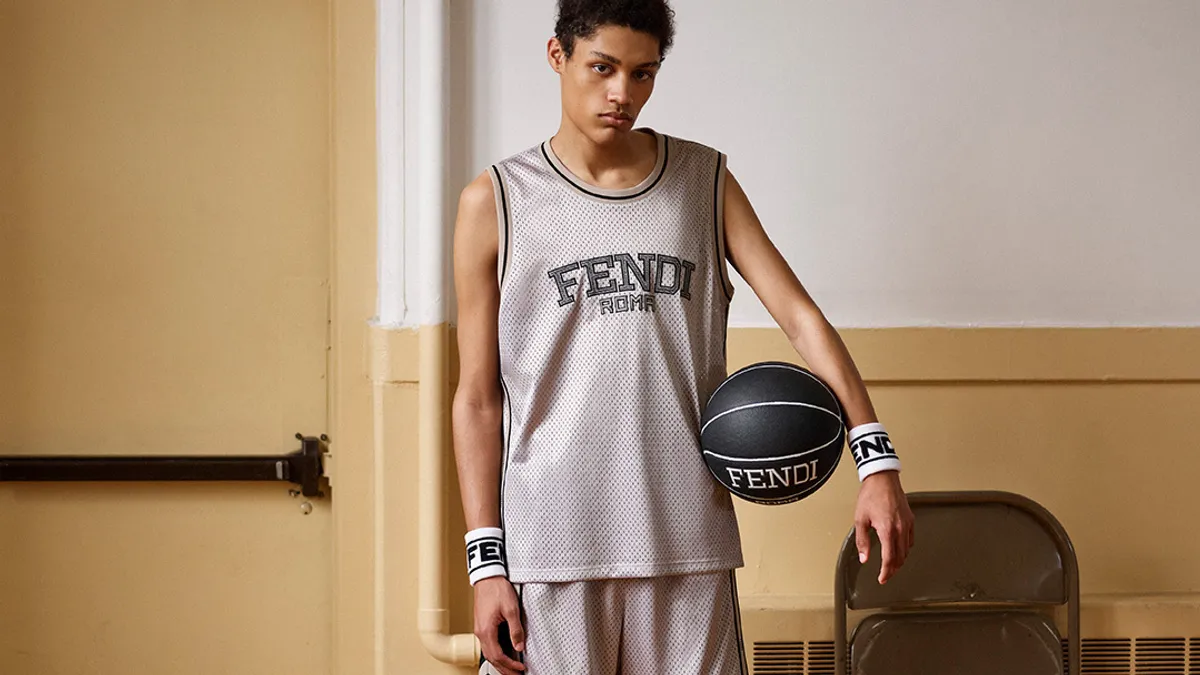 A man standing on a basketball court near the door wears a Fendi logo jersey, shorts, socks and sneakers, and hods a fendi logo basketball.