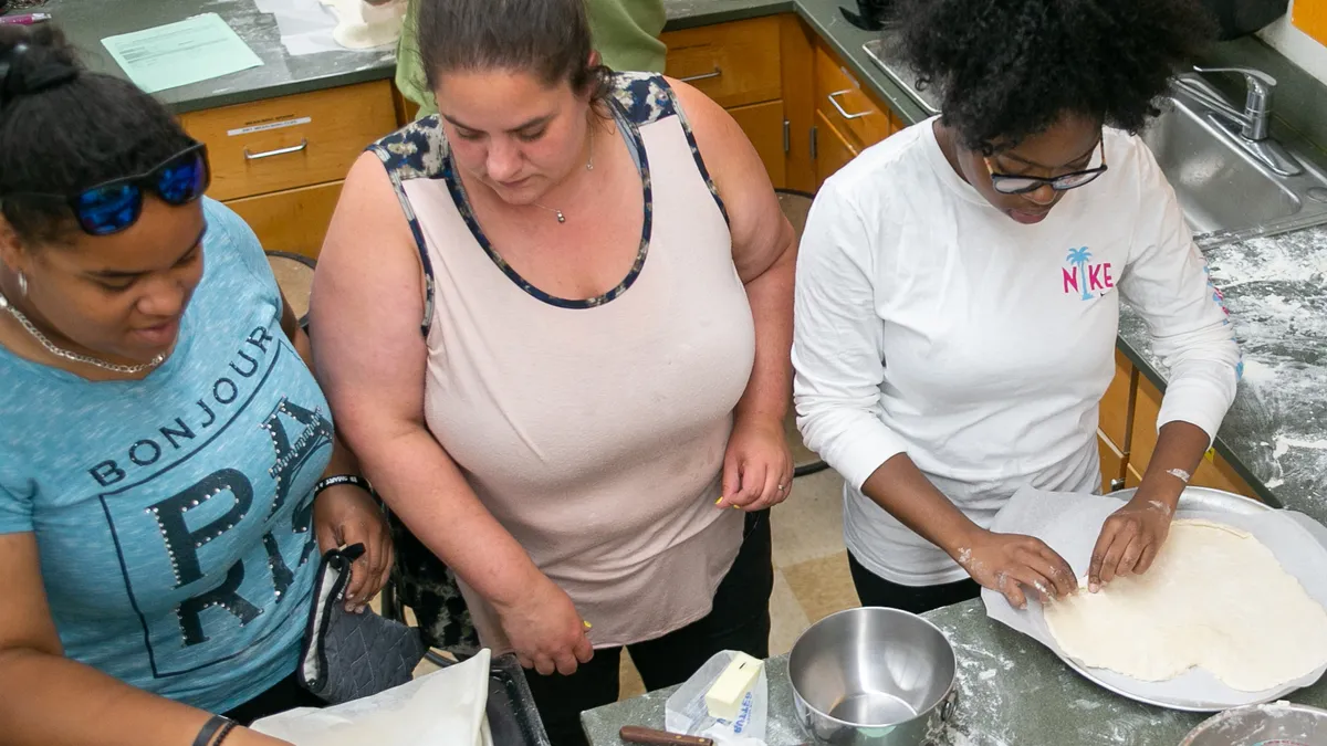 Three students stand side-by-side and are working with dough with their hands in a kitchen. The photo angle is from above looking down and across at the students.