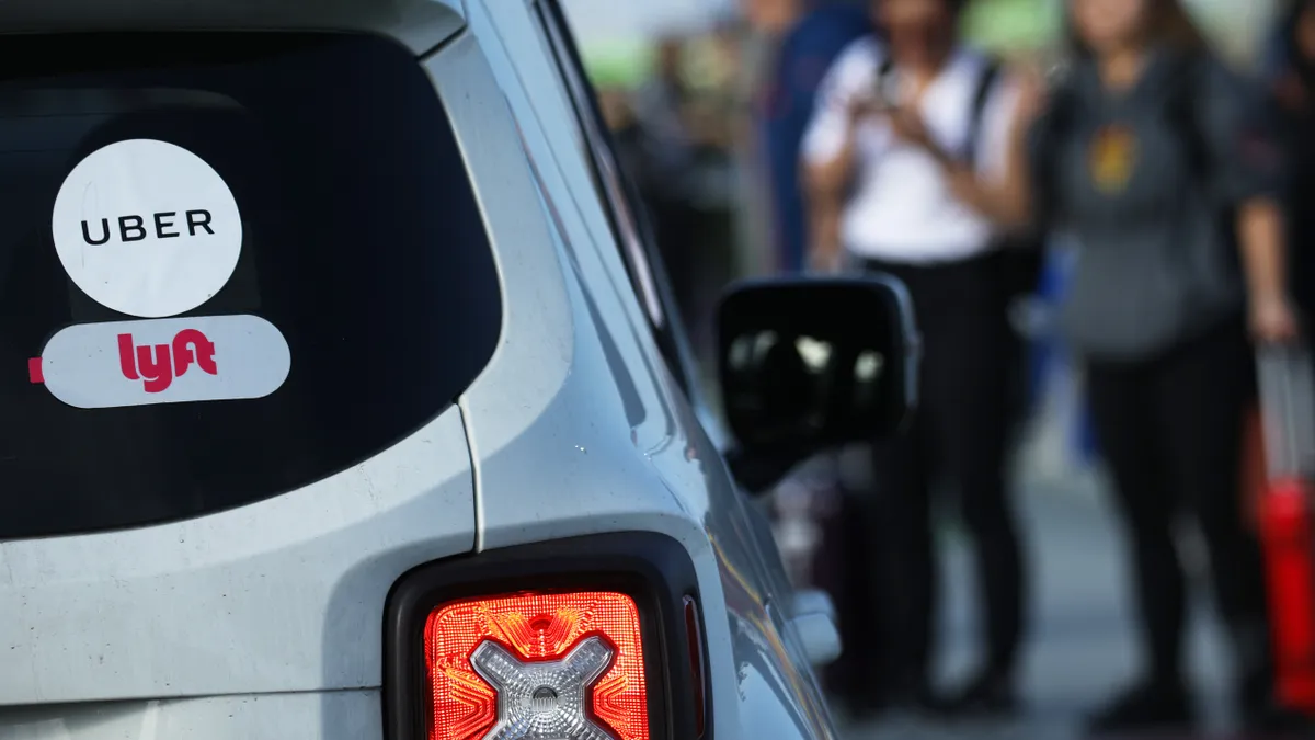 Close-up of the right rear of a small SUV with Uber and Lyft stickers on the rear window, with a blurred group of people standing to the right.