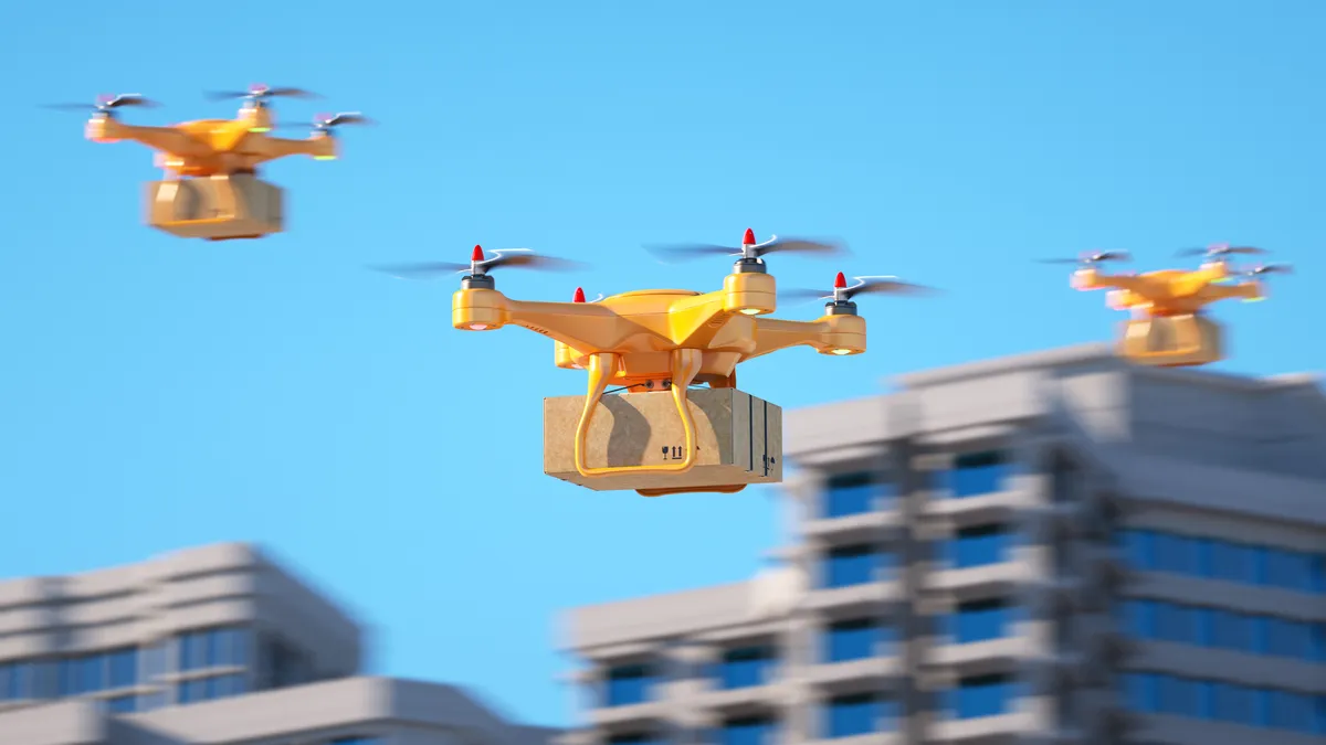 Three yellow delivery drones carrying packages fly near several tall buildings.