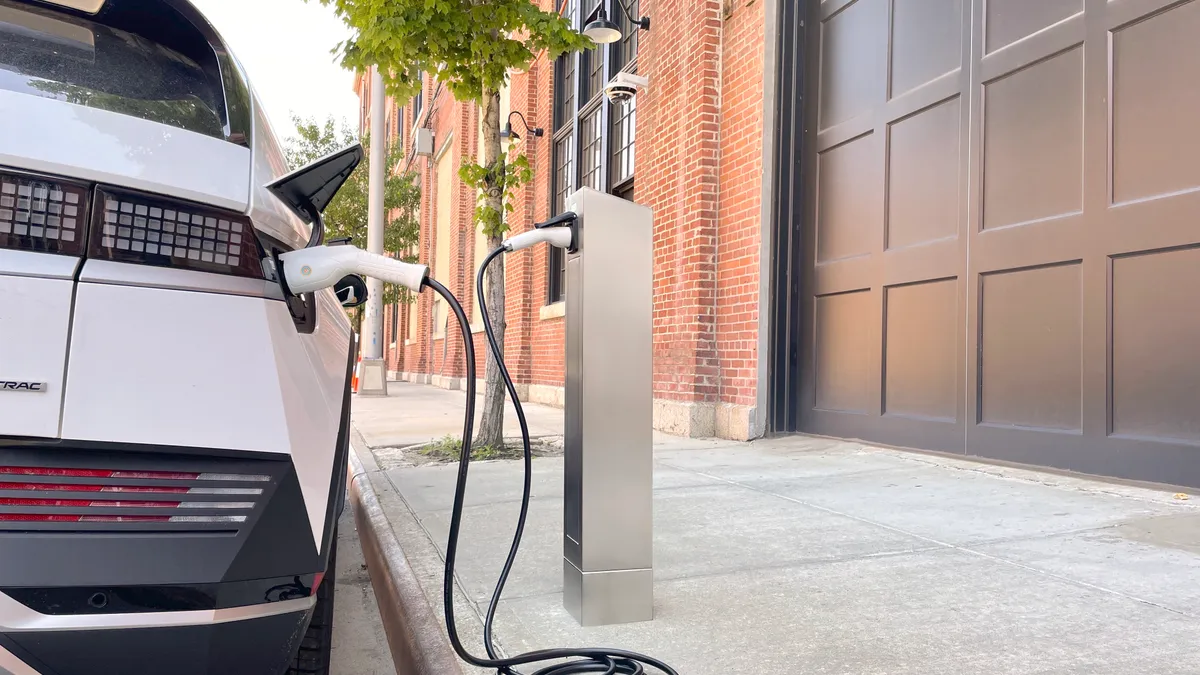 The right rear corner of a white car is parked on a street plugged in to an electric vehicle charging post.