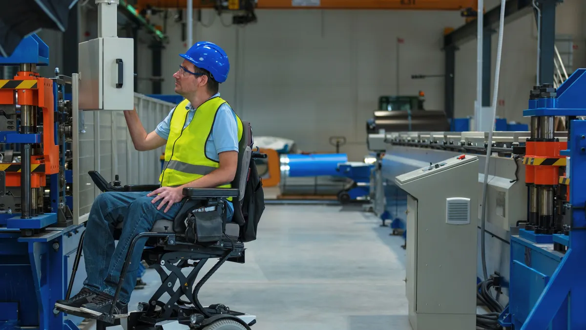 A person in a wheelchair and wearing a blue hardhat and yellow vest, operating a machine in a factory.