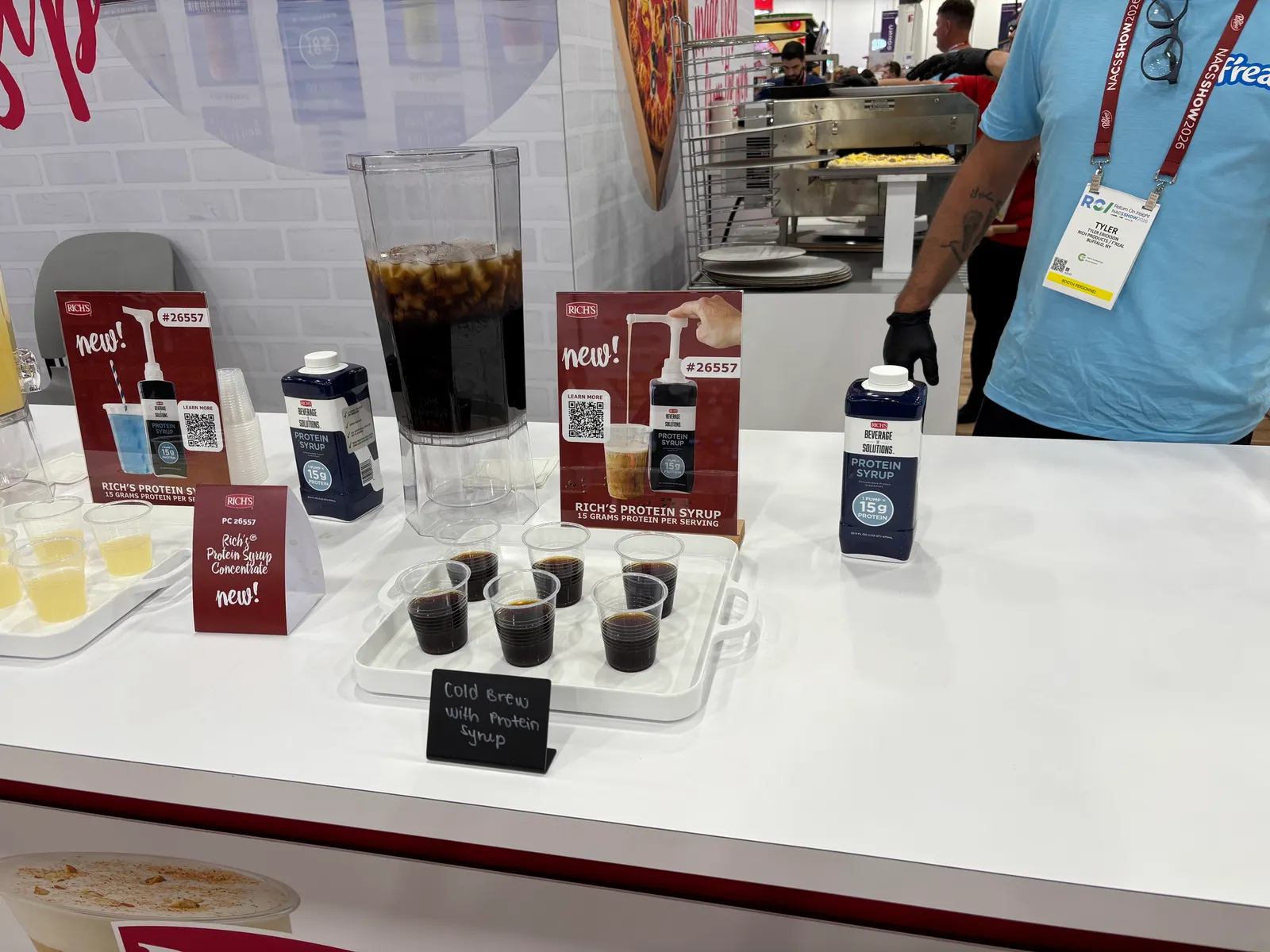 A display with a container, a pump, several cups and signs. The signs say New! Rich's protein syrup. 15 grams of protein per serving.