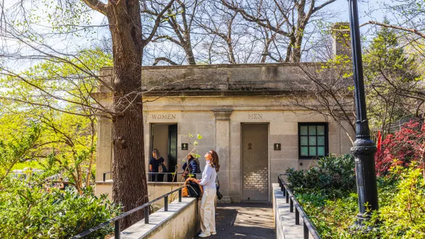 People stand near a stone building with two doors with the words Women and Men above them surrounded by spring foliage.