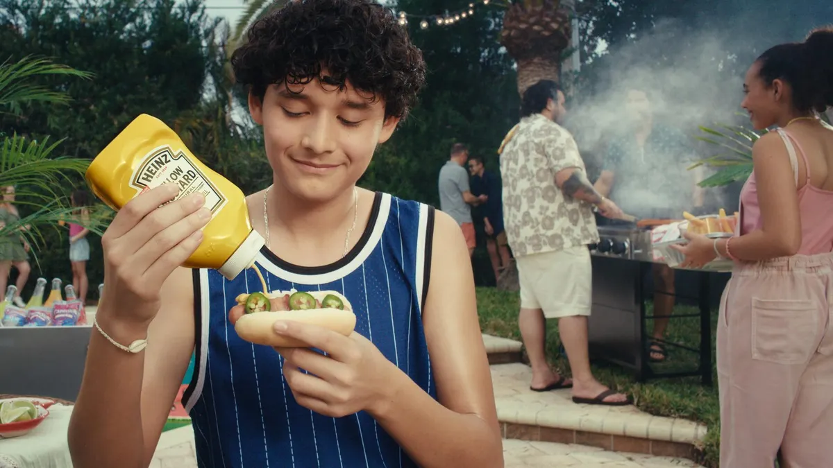 A young cookout attendee in a sports jersey squirts Heinz yellow mustard on a hot dog