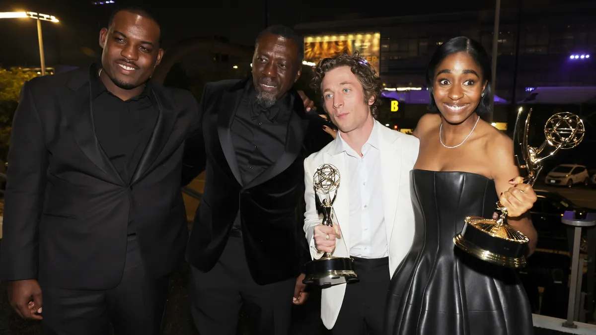 Two people in black tuxedos, one person in a white dinner jacket and white shirt, and a perosn in a black leather dress stand holding Emmy Awards.