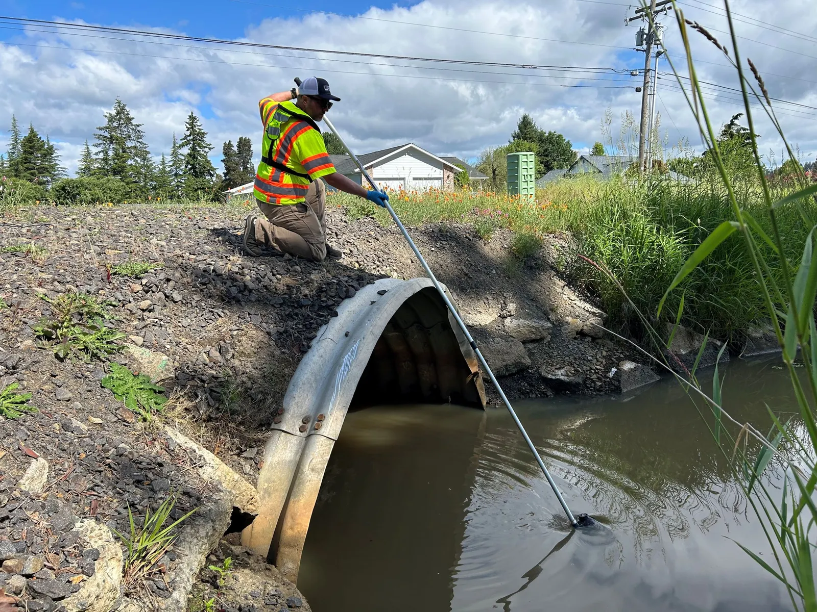A person on top of a large ditch pipe dips a long pole into a pool of water in the ditch below.