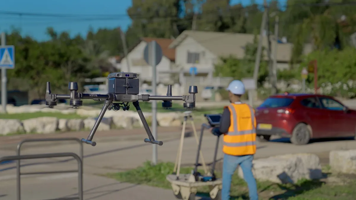 A drone floats in the foreground while a slightly blurry worker with a hardhat on in the background works on a tablet.