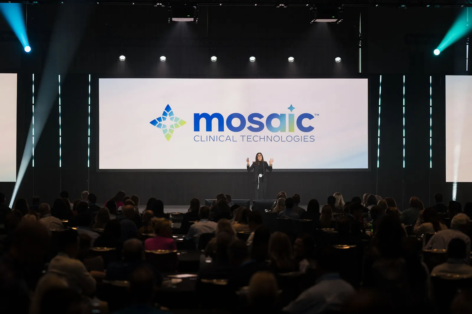 A person speaks on a stage in front of a large screen, which says &quot;Mosaic clinical technologies&quot;