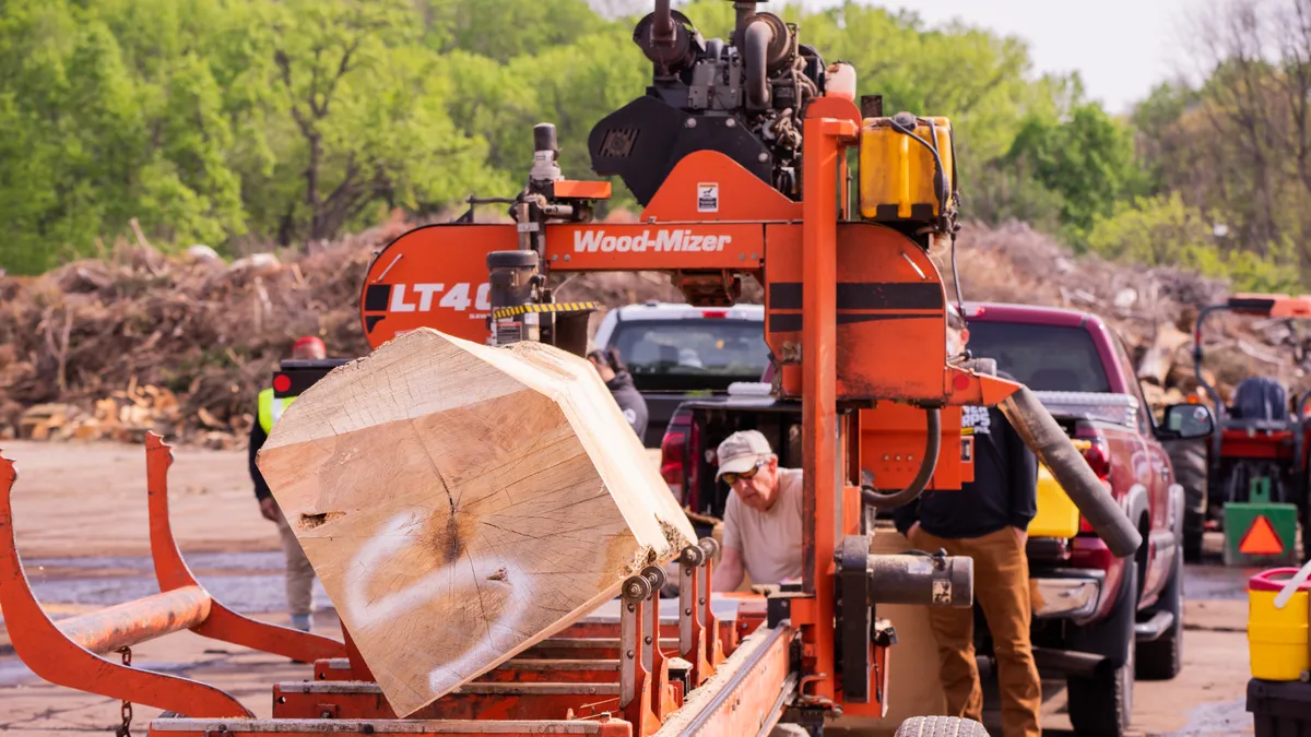 A piece of wood sits on equipment.  A person wearing a hat looks through the equipment. Behind the person is a pile of branches and trucks.