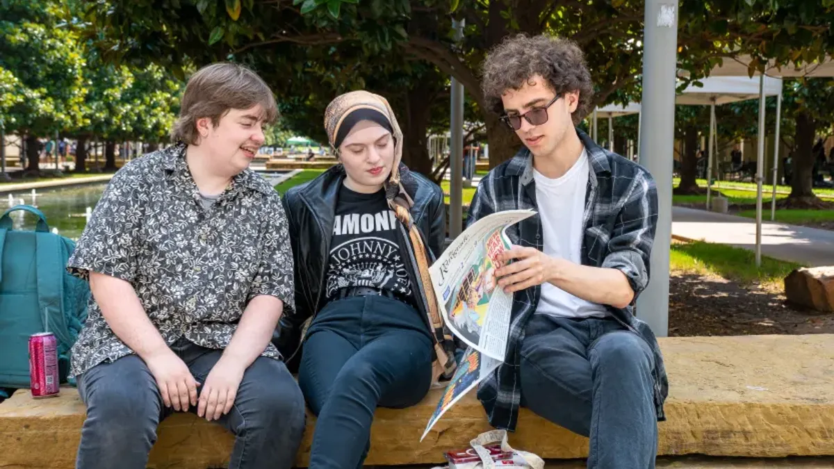 Three students sit at a bench flipping through a newspaper.